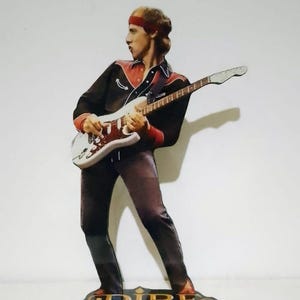 Puede incluir: Recorte de madera de un guitarrista con una camisa negra con detalles rojos, pantalones marrones y una diadema roja, tocando una guitarra blanca. La base muestra las palabras "Dire Straits" en letras doradas sobre un fondo negro.
