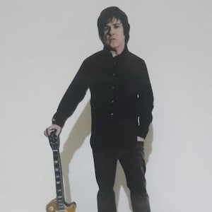 以下が含まれることがあります： ギターを持ったゲイリー・ムーアの段ボール製切り抜き。黒いシャツとズボンを着用しています。ベースは、赤と白で「GARY MOORE」という名前が入った黒い三角形です。