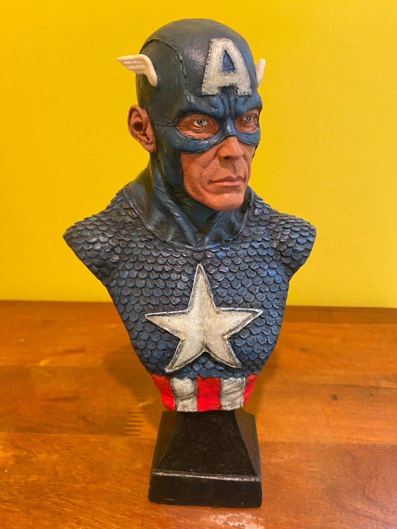 Puede incluir: Busto detallado de Capit&aacute;n Am&eacute;rica, con un casco azul con una "A" blanca y alas, una m&aacute;scara azul y un pecho estrellado. La escultura est&aacute; montada sobre una base negra, mostrando la imagen del ic&oacute;nico superh&eacute;roe.
