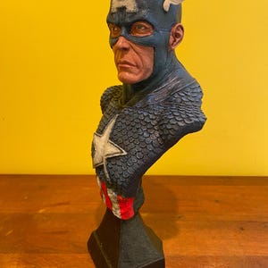 Puede incluir: Busto detallado del Capit&aacute;n Am&eacute;rica, con un casco azul con una "A" blanca y alas. El busto tiene un esquema de color azul y rojo, con una estrella blanca en el pecho. La escultura est&aacute; montada sobre una base negra.