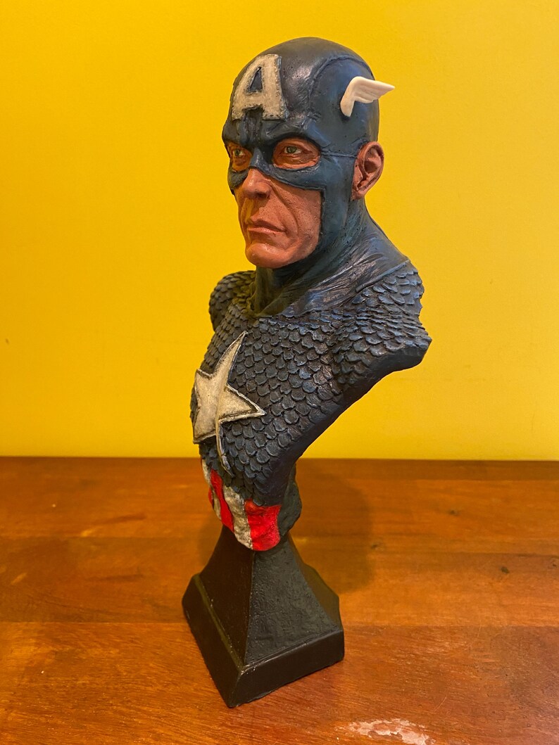 Puede incluir: Busto detallado del Capit&aacute;n Am&eacute;rica, con un casco azul con una "A" blanca y alas. El busto tiene un esquema de color azul y rojo, con una estrella blanca en el pecho. La escultura est&aacute; montada sobre una base negra.