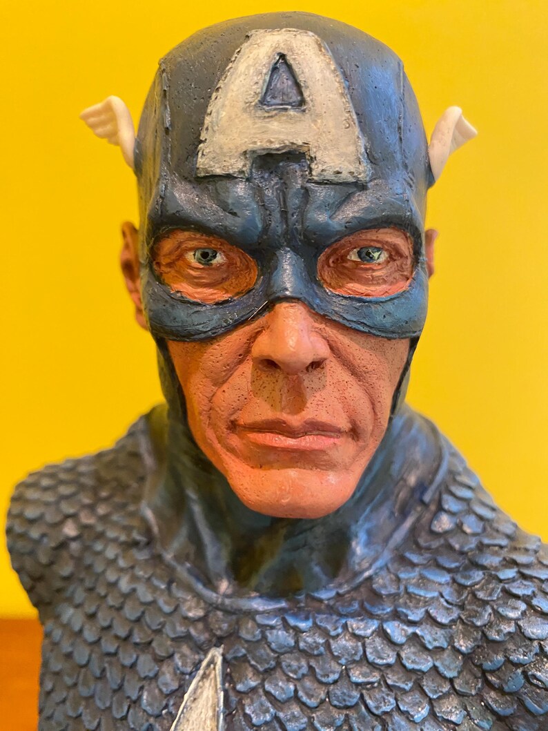 Puede incluir: Busto detallado de Capit&aacute;n Am&eacute;rica, con un casco azul con una "A" blanca y detalles alados. El busto muestra la m&aacute;scara del personaje, sus rasgos faciales y un dise&ntilde;o de armadura texturizada en forma de escamas. La paleta de colores incluye tonos de azul, rojo y blanco.