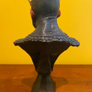 Puede incluir: Busto verde oscuro de una criatura con peque&ntilde;os cuernos y orejas puntiagudas. La escultura tiene una superficie texturizada y un cuello detallado con escamas. El busto est&aacute; montado sobre una base negra geom&eacute;trica, sobre un fondo amarillo.