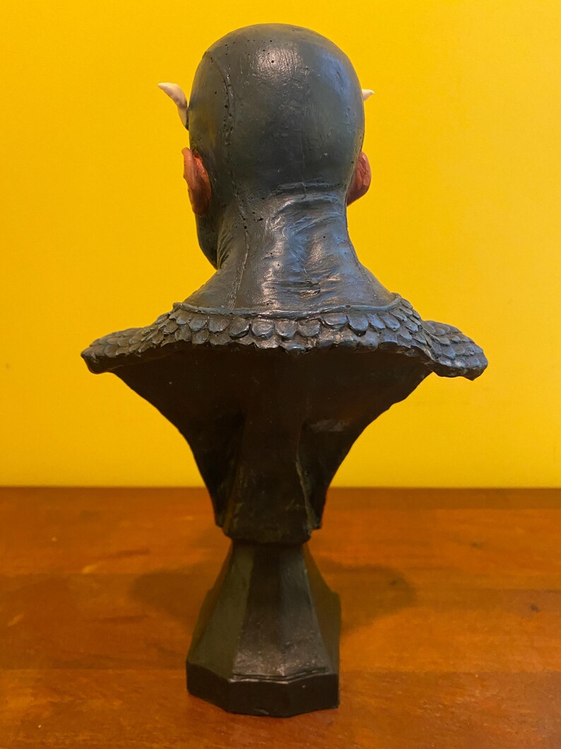 Puede incluir: Busto verde oscuro de una criatura con peque&ntilde;os cuernos y orejas puntiagudas. La escultura tiene una superficie texturizada y un cuello detallado con escamas. El busto est&aacute; montado sobre una base negra geom&eacute;trica, sobre un fondo amarillo.