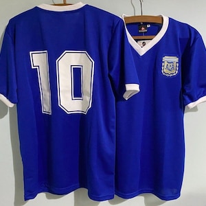 Pode incluir: Duas camisas de futebol azuis. Uma camisa tem o número 10 nas costas em branco com contorno branco. A outra camisa tem decote em V, o emblema da seleção argentina e acabamento branco nas mangas e gola.