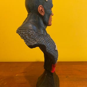 Puede incluir: Busto de Capit&aacute;n Am&eacute;rica, con casco azul y detalles blancos, una estrella roja y blanca en el pecho y una base negra. La escultura presenta un acabado texturizado y rasgos faciales realistas.