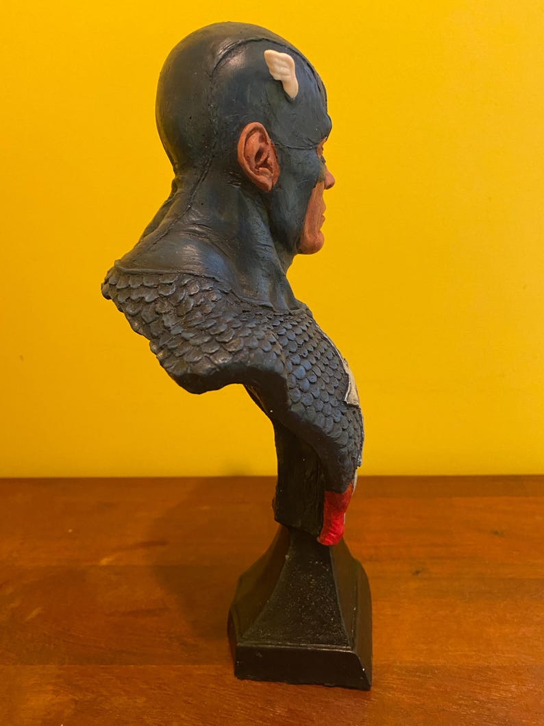 Puede incluir: Busto de Capit&aacute;n Am&eacute;rica, con casco azul y detalles blancos, una estrella roja y blanca en el pecho y una base negra. La escultura presenta un acabado texturizado y rasgos faciales realistas.