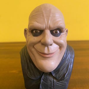 May include: A bust of Uncle Fester from The Addams Family. The figure has a bald head, dark eyes, and a wide, smiling mouth. He wears a black collared shirt. The bust is made of a light beige material.