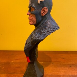 Puede incluir: Un busto de Capit&aacute;n Am&eacute;rica, con un casco azul con un emblema de ala blanca. La escultura tiene un rostro detallado y un traje azul y rojo. El busto est&aacute; montado sobre una base negra, mostrando un dise&ntilde;o cl&aacute;sico de superh&eacute;roe.