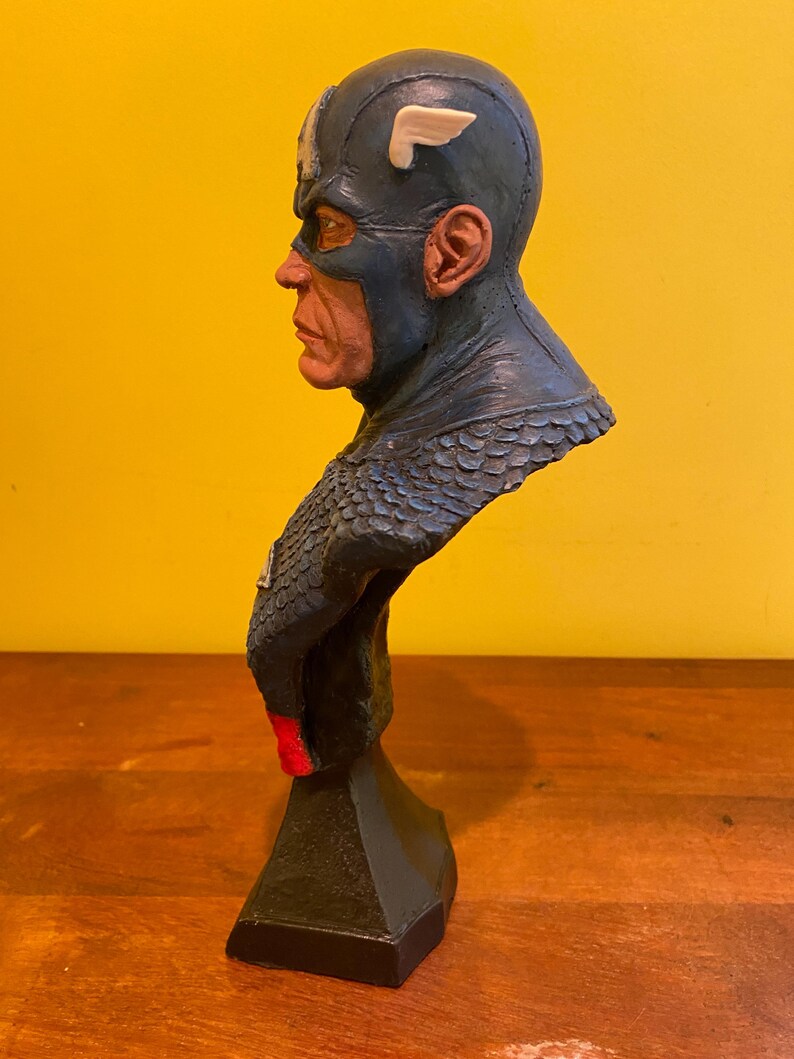 Puede incluir: Un busto de Capit&aacute;n Am&eacute;rica, con un casco azul con un emblema de ala blanca. La escultura tiene un rostro detallado y un traje azul y rojo. El busto est&aacute; montado sobre una base negra, mostrando un dise&ntilde;o cl&aacute;sico de superh&eacute;roe.