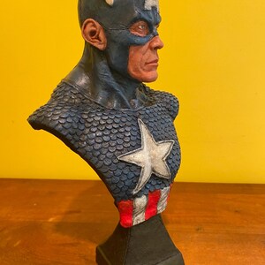 Puede incluir: Busto de Capit&aacute;n Am&eacute;rica, con un casco azul con una 'A' blanca y un emblema de ala. El pecho muestra una estrella blanca, y la base tiene rayas rojas y blancas. La escultura est&aacute; sobre una base negra.