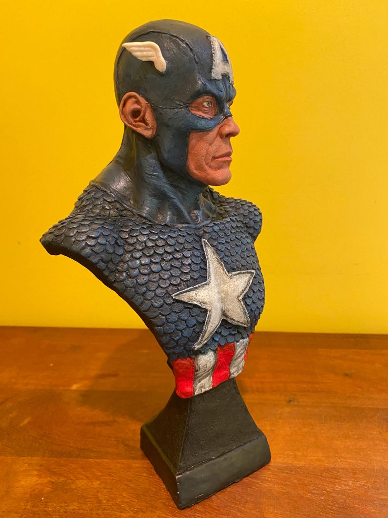 Puede incluir: Busto de Capit&aacute;n Am&eacute;rica, con un casco azul con una 'A' blanca y un emblema de ala. El pecho muestra una estrella blanca, y la base tiene rayas rojas y blancas. La escultura est&aacute; sobre una base negra.