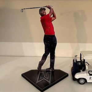 May include: A black diamond-shaped base with a silver logo supports a figure of a golfer in a red shirt and black pants mid-swing. A white golf cart with black wheels is to the right.