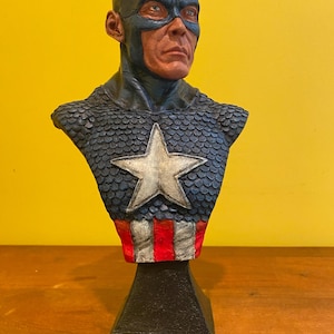 Puede incluir: Busto de Capit&aacute;n Am&eacute;rica. El busto presenta un casco azul con una "A" blanca y alas. El pecho tiene una estrella azul y plateada, con rayas rojas y blancas en la base. La escultura est&aacute; sobre una base negra.
