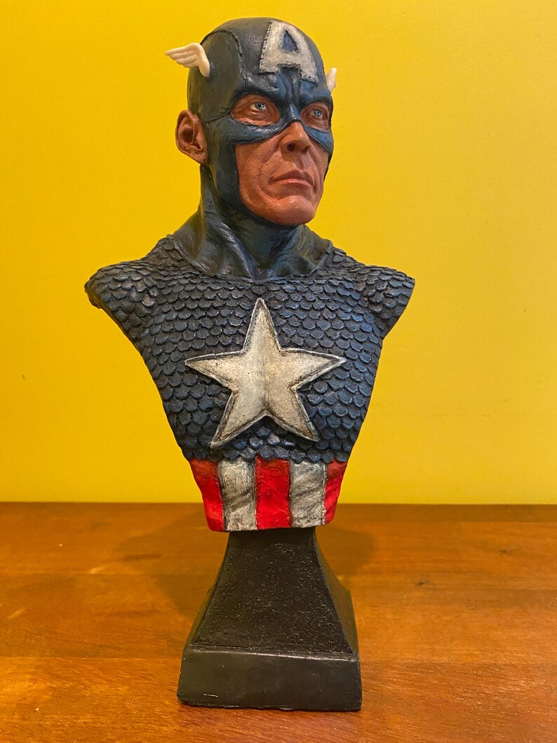 Puede incluir: Busto de Capit&aacute;n Am&eacute;rica. El busto presenta un casco azul con una "A" blanca y alas. El pecho tiene una estrella azul y plateada, con rayas rojas y blancas en la base. La escultura est&aacute; sobre una base negra.
