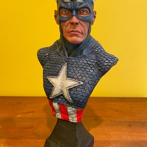 Puede incluir: Busto detallado de Capit&aacute;n Am&eacute;rica, con un casco azul con una "A" blanca y alas, una m&aacute;scara azul y un emblema en forma de estrella en el pecho. La escultura tiene una apariencia texturizada, similar a escamas, en los hombros y una secci&oacute;n a rayas rojas y blancas en la base.