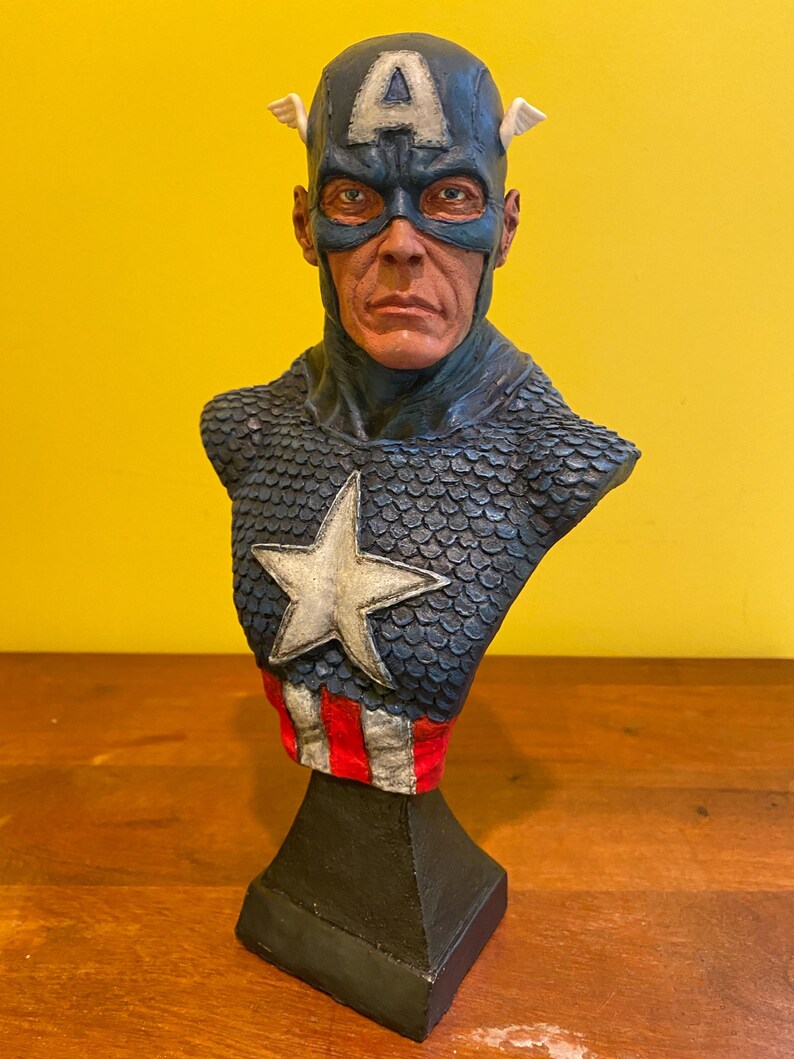 Puede incluir: Busto detallado de Capit&aacute;n Am&eacute;rica, con un casco azul con una "A" blanca y alas, una m&aacute;scara azul y un emblema en forma de estrella en el pecho. La escultura tiene una apariencia texturizada, similar a escamas, en los hombros y una secci&oacute;n a rayas rojas y blancas en la base.
