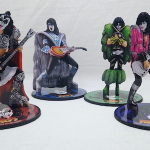Puede incluir: Cuatro figuras decorativas de la banda de rock KISS, cada una con un disfraz y una pose diferentes. Las figuras están sobre bases circulares y sostienen guitarras. Las figuras están pintadas con colores vibrantes y diseños detallados.