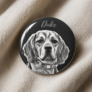 May include: A circular badge with a black background, displaying a detailed monochrome illustration of a beagle. The dog, wearing a collar, is the central focus. The name "Duke" is written above the dog in a cursive font. The badge rests on a neutral fabric.