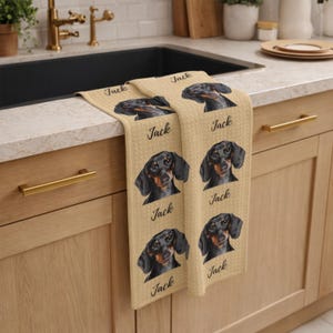 May include: Beige kitchen towel featuring a repeating pattern of a black and brown dachshund dog portrait and the name "Jack" in a script font. The towel is folded over a kitchen sink.