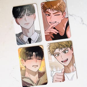 Può includere: Quattro carte collezionabili rettangolari con illustrazioni di personaggi in stile anime. Due carte mostrano individui con capelli scuri, una mostra un personaggio biondo che fa l'occhiolino e un'altra un personaggio biondo che sorride col pollice in su.
