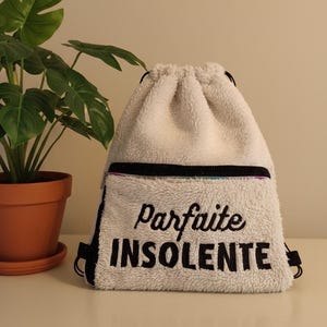 Peut inclure: Un sac à cordon blanc et duveteux avec les mots "Parfaite Insolente" brodés en noir. Le sac a une poche zippée noire et des cordons noirs. Une plante en pot est en arrière-plan.