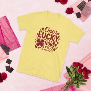 Może przedstawiać: Jasnożółty t-shirt z krótkim rękawem z ciemnobordowym nadrukiem graficznym o treści One LUCKY NURSE. Wzór zawiera czterolistną koniczynę, stetoskop w kształcie serca, małe kwiaty, liście oraz ikony związane z tematyką medyczną.