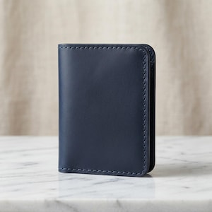 May include: A vertical navy blue leather card holder featuring a smooth matte finish and visible tonal stitching along the rounded edges. The wallet has a slim, rectangular construction with a clean, minimalist design and a single visible compartment.
