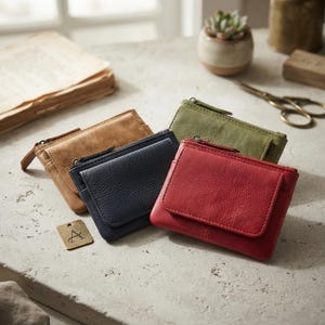 May include: Four small leather wallets in red, navy blue, olive green, and tan. Each wallet features a zipped top closure and a front flap pocket. A small rectangular metal tag with the letter A engraved on it sits next to the wallets.