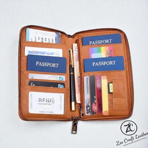 Könnte beinhalten: Offene, hellbraune Reisebrieftasche aus Leder mit mehreren Fächern für Pässe, Kreditkarten und Bargeld. Enthält einen schwarzen Stift und einen Reißverschluss. Die Brieftasche ist mit "RFID Protected" gekennzeichnet und trägt den Text "Zee Craft Leather".