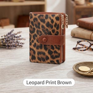 May include: A bi-fold wallet featuring a brown and black leopard print pattern. The wallet includes a brown leather-like strap with a snap button closure and two gold-tone metal zippers along the side. The text Leopard Print Brown is visible.