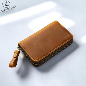 Puede incluir: Una cartera rectangular de cuero marrón con cierre de cremallera. La cartera tiene una lengüeta y una cremallera marrón oscuro. El cuero tiene un aspecto envejecido. La cartera es de tamaño compacto, adecuada para llevar tarjetas y dinero en efectivo.