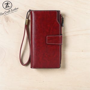 May include: A burgundy leather wallet with a wrist strap. The wallet has a snap closure and a zip. The Zee Craft Leather logo is in the upper left corner. The wallet is on a light wood surface.