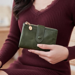 Puede incluir: Una cartera plegable de color verde oliva oscuro con acabado liso, cremallera metálica dorada con un pequeño dije ovalado y una solapa con cierre de botón a presión en la parte frontal. La cartera tiene una forma rectangular y compacta.