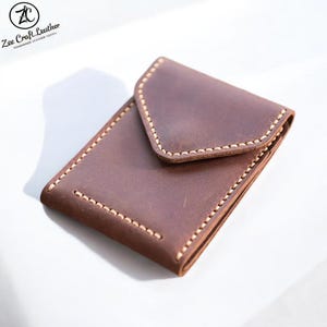 May include: A brown leather wallet with a flap closure. The rectangular wallet has a stitched border with light-coloured thread. The Zee Craft Leather logo is in the upper left corner.