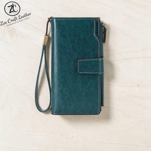 May include: A dark teal wallet with a textured finish, featuring a matching wrist strap, a snap-button closure, and a side zipper. The wallet includes white contrast stitching along the edges and a logo in the corner reading Zee Craft Leather.