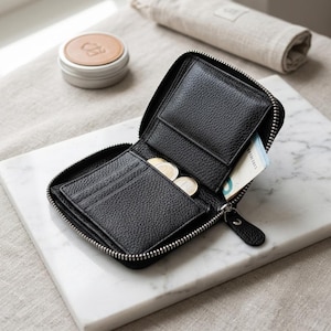 May include: A black pebbled leather wallet with a silver zip closure. The wallet is open, revealing multiple card slots, a central compartment containing two round metal coins, and a side pocket holding a piece of paper currency.