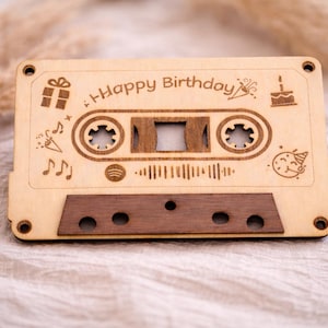 Könnte beinhalten: Eine Holzkassette mit der Gravur "Happy Birthday". Das Design umfasst Noten, ein Geschenk, einen Kuchen und eine Partyhütchen. Die Kassette hat einen dunkelbraunen unteren und einen hellbraunen oberen Teil.