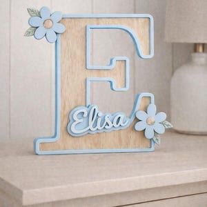 Könnte beinhalten: Holzbuchstabe "E" mit hellblauer Umrandung und dem Namen "Elisa" in Schreibschrift. Der Buchstabe ist mit hellblauen Blumen mit goldenen Zentren und grünen Blättern verziert. Der Holzbuchstabe steht auf einer hellen Oberfläche.