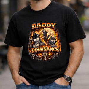 May include: Black t-shirt with a graphic design. The design features three wolves, flames, and the words "DADDY DOMINANCE." One wolf wears a hat. A full moon is in the background, and a skull is at the bottom.