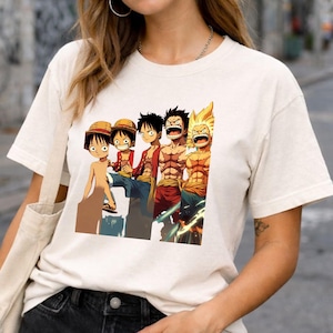 Może przedstawiać: Kremowa koszulka z kolorową grafiką przedstawiającą pięć postaci z anime w różnych pozach. Postacie są przedstawione ze szczegółowymi wyrazami twarzy i strojami. Koszulka jest wykonana z miękkiej, jasnej tkaniny.