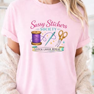 Può includere: T-shirt rosa chiaro con la scritta "Sassy Stitchers Society" e una grafica di strumenti di cucito. La grafica include una bobina di filo viola, un ago e forbici dorate. Il testo "STITCH. LAUGH. REPEAT." è sotto.