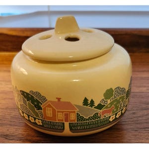 May include: A cream-colored ceramic jar with a matching lid featuring three circular holes. The exterior is decorated with a colorful wrap-around illustration of a small house, green rolling hills, trees, a brown horse, and yellow flowers.