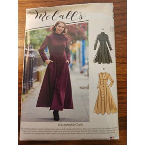 May include: A McCall's sewing pattern envelope featuring a long-sleeved, button-front dress with a high neckline and pockets. The cover displays a model wearing a deep burgundy version, alongside line drawings of two variations in black and tan.