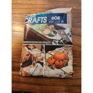 Può includere: Una busta per cartamodello McCall's Crafts, numero 608, per centrotavola e decorazioni festive. La confezione presenta illustrazioni fotografiche di tavoli da pranzo decorati con zucche, vegetazione e stoviglie su uno sfondo scuro.