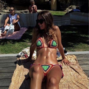 Puede incluir: Un conjunto de bikini de ganchillo hecho a mano en rojo, verde y morado. La parte superior presenta copas triangulares con centros verdes y bordes rojos. La parte inferior tiene un diseño a juego. El bikini se usa al aire libre en un día soleado.