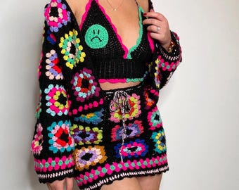 Puede incluir: Un conjunto de tres piezas de ganchillo que incluye un bralette negro con un parche verde de cara triste, un cárdigan de manga larga a juego y una minifalda. Las prendas están hechas de hilo negro con motivos cuadrados coloridos en rosa, azul, amarillo y morado.