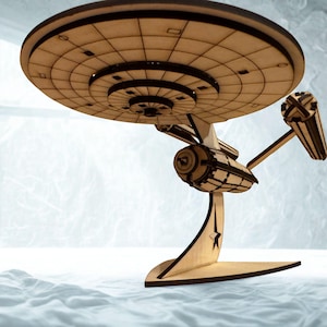 May include: A wooden model spaceship, with a circular main section and intricate engine details. Constructed from light brown wood, it rests on a matching base. The design is evocative of a classic science fiction starship, suitable for display.