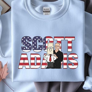 May include: Pale blue sweatshirt with the name "SCOTT ADAMS" in a patriotic design. The letters "SCOTT" are filled with a stars and stripes pattern, while "ADAMS" has red and white stripes. A cartoon image of a man is on the sweatshirt.
