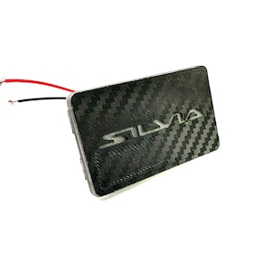 Pode incluir: Um painel retangular de fibra de carbono preta com a palavra "SILVIA" em letras prateadas. O painel tem uma superfície texturizada e dois fios, um vermelho e outro preto, que se estendem dele.